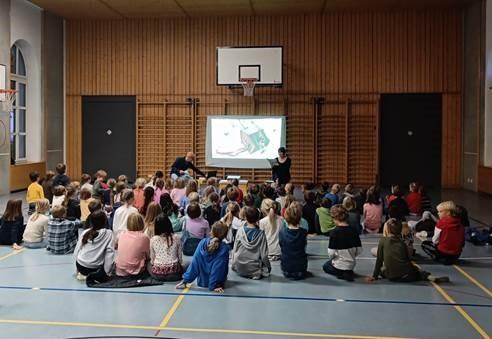 Kinder sitzen in der Turnhalle am Boden und schauen den Darbietungen zu
