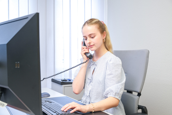 Eine Auszubildende, die im Büro an einem Computer sitzt und telefoniert.
