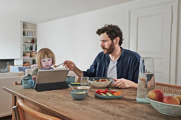 Kleinkind vor dem Laptop, Vater versucht es vergeblich zu füttern.