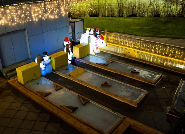 Nachtansicht einer beleuchteten Eisminigolf-Anlage mit mehreren parallelen Bahnen. Die Bahnen sind mit Holzrahmen eingefasst und mit einer eisähnlichen Oberfläche belegt. Am Ende jeder Bahn stehen beleuchtete Winterfiguren: Schneemänner mit Mützen und Schals, Pinguine und ein grosser Eisbär. Entlang der rechten Wand hängen Lichterketten, die warmes Licht abgeben. Im Hintergrund ist eine Grünfläche mit Bäumen zu erkennen. Die Anlage ist festlich dekoriert für die Winter- und Weihnachtszeit.