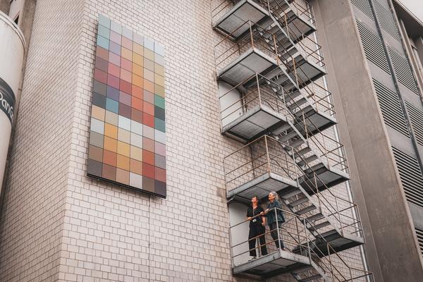 Zwei Frauen schauen von einer Treppe aus auf farbige Solarpanels an der Wand.